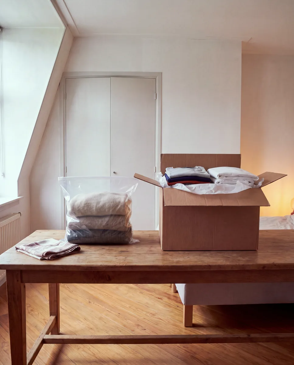 Gevouwen wollen truien in een dichte doorzichtige zak naast een open verhuisdoos met katoenen kleding tussen vloeipapier, op een houten tafel bij het raam.