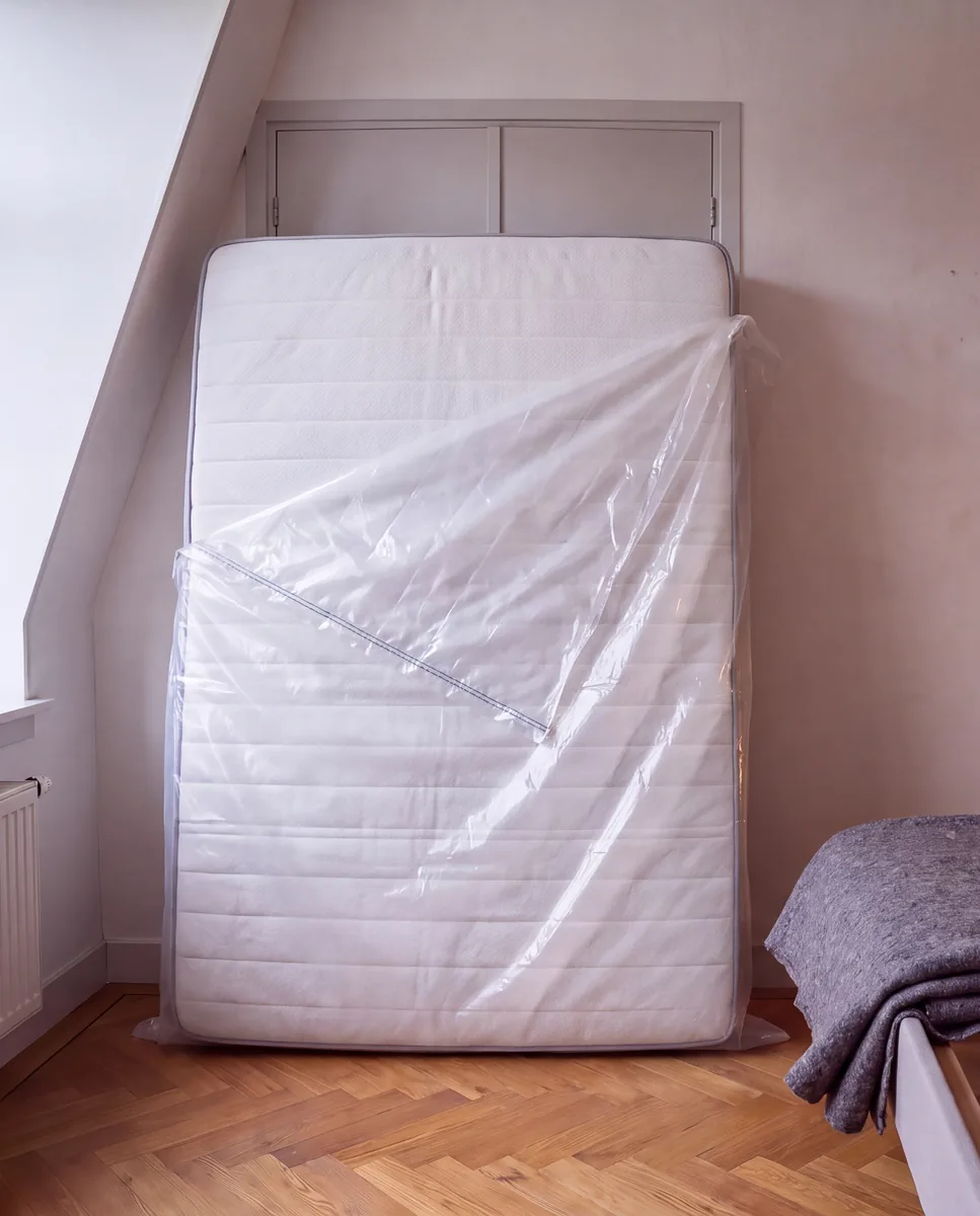 Kaal matras rechtop naast het bedframe, half in een doorzichtige matrashoes, met een opgevouwen verhuisdeken op de lattenbodem.