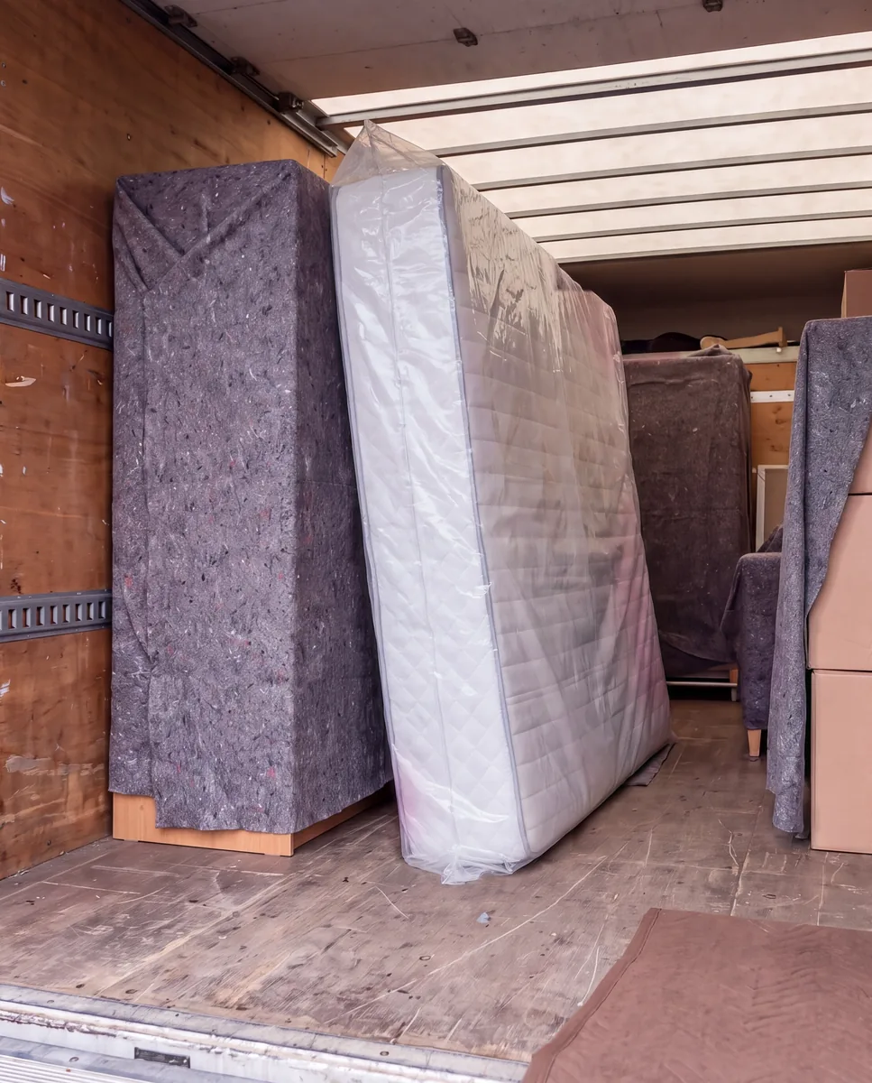 Matras in een doorzichtige verhuismatrashoes staat rechtop in de laadruimte van de verhuiswagen, tussen een ingedekende kast en verhuisdozen met een verhuisdeken ertussen.