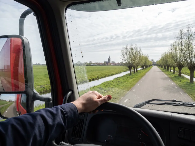 Uitzicht vanuit de cabine van de verhuiswagen op een rechte polderweg met knotwilgen en een kerktoren aan de horizon